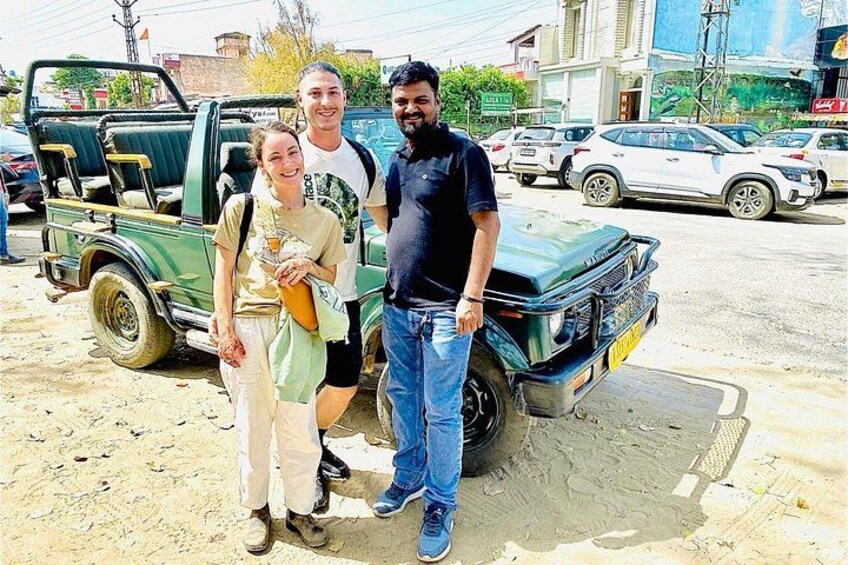 Three people standing in front of a green pickup truck on a dusty street with buildings and parked cars in the background. 1