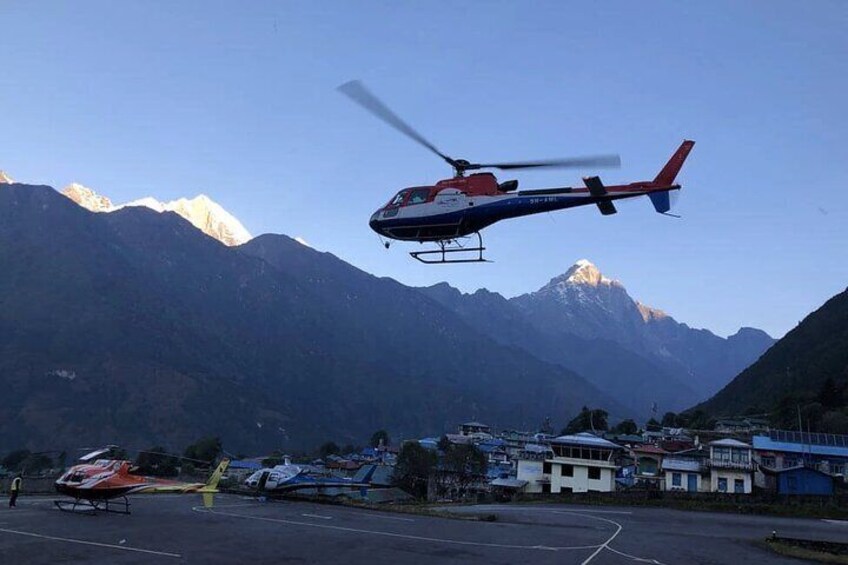A helicopter flying over a mountainous area with a helipad and buildings in the foreground. 2