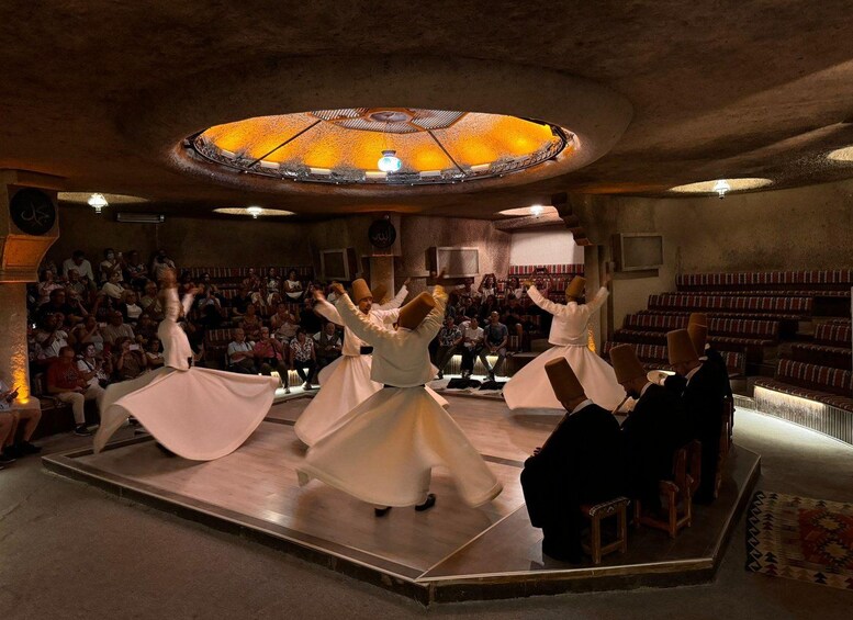 A dance performance with dancers in white on a stage, an audience in the background, and a circular ceiling light. 2