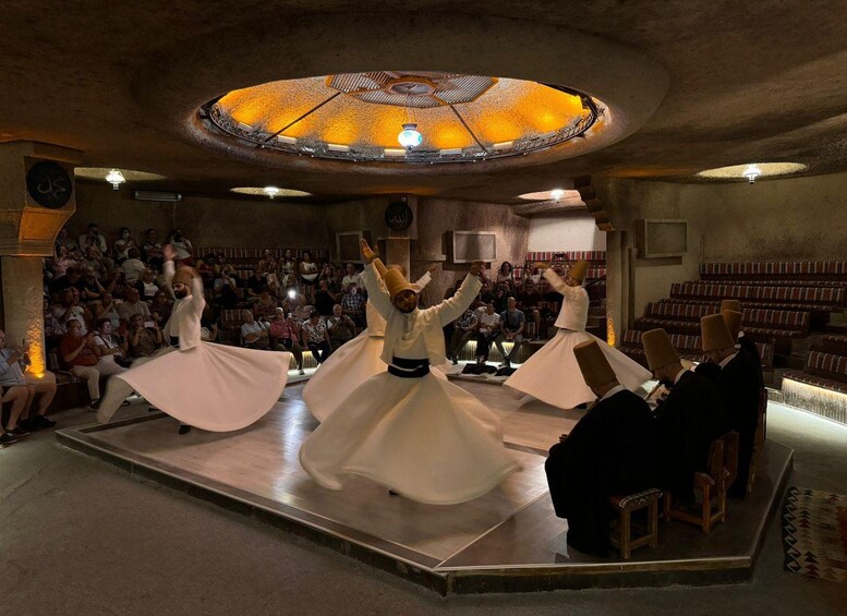 A cultural performance with dancers in white attire on a stage, an audience seated in the background, and a unique circular ceiling design. 5