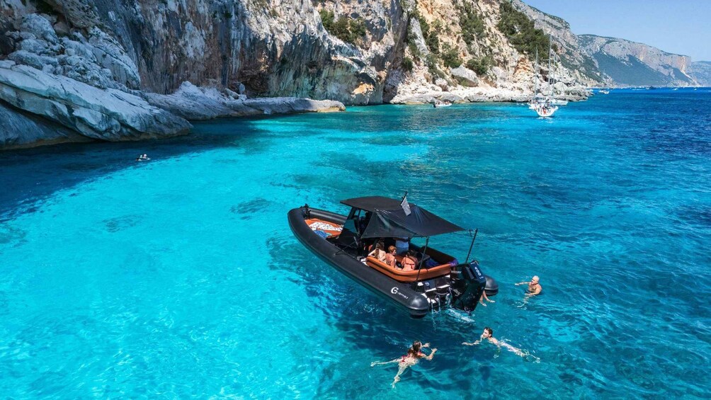 Ein Boot in klarem, blauem Wasser nahe schroffer Felsen, während Menschen schwimmen. 5