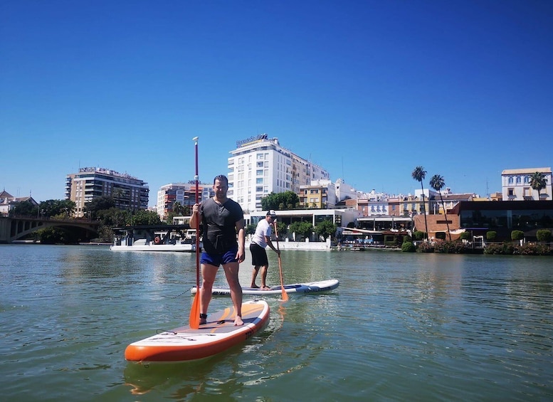 Persone che fanno paddleboarding su un fiume tranquillo, con edifici e palme sullo sfondo. 5