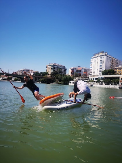 Due persone fanno paddleboarding su un fiume calmo, con edifici sullo sfondo. 2