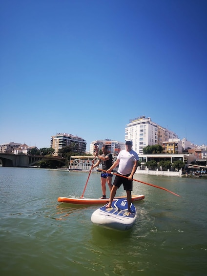 Due persone che fanno paddleboard su un fiume, con edifici sullo sfondo. 4
