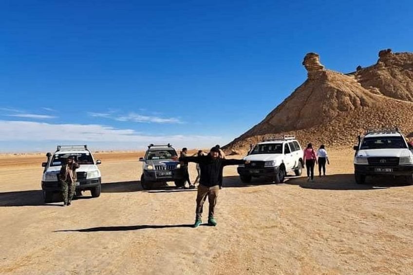 A desert landscape with a large rock formation, several vehicles, and people. 3