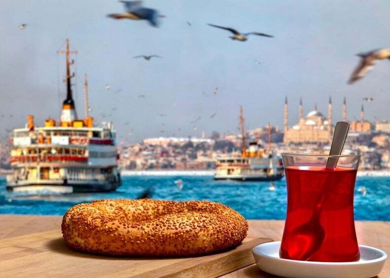 A wooden table with a sesame seed bagel and a glass of red tea, with a cityscape and boats in the background. 2