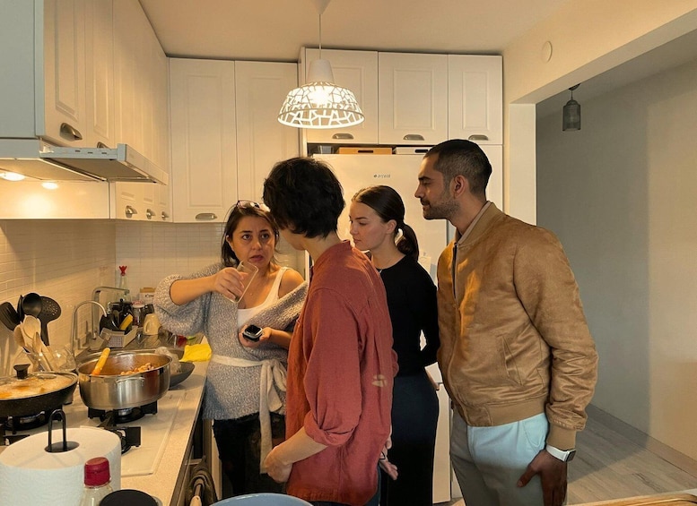 A group of people in a modern kitchen with white cabinets, a hanging light fixture, and various cooking utensils on the counter. 1