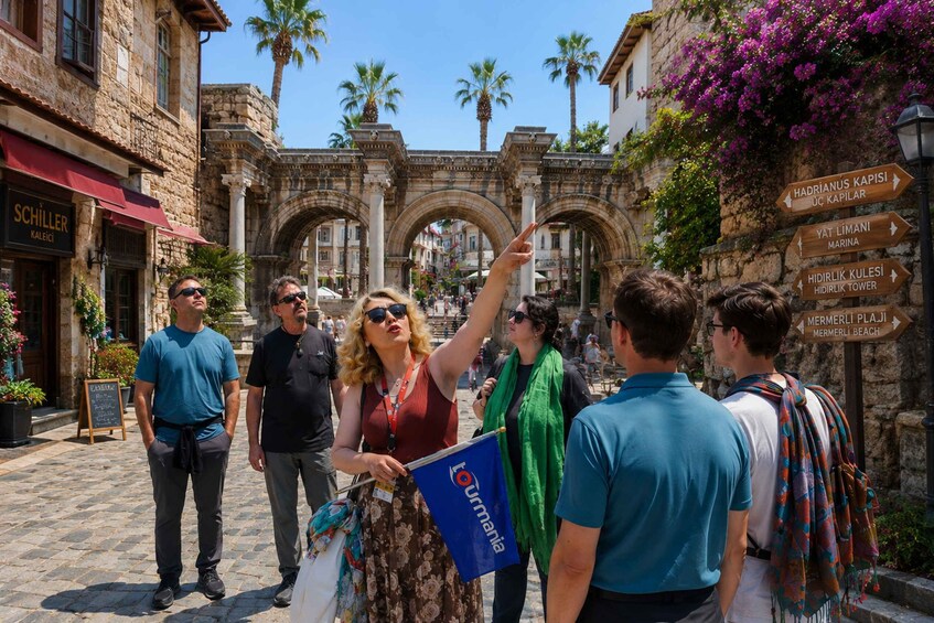 A group of people in a historic stone-paved street with arches, a sign pointing to Hadrian's Gate, and a blue flag with 'Tourania' on it. 3