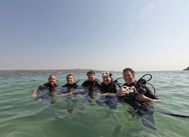 Four people in a clear blue sea, wearing scuba diving gear, with a distant landmass and a clear sky. 3