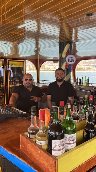 Two men behind a bar with various bottles of alcohol, a DJ controller, and a sign that reads 'FRENCH BLUE'. 5