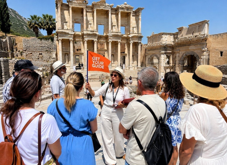 Des touristes visitent un site archéologique antique, avec ses colonnes et ses arcades. Un guide tient une pancarte. Le ciel est d’un bleu limpide. 2