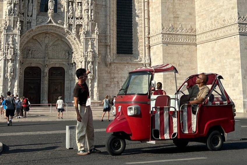 A red three-wheeled vehicle parked in front of a historic building with intricate carvings. 1