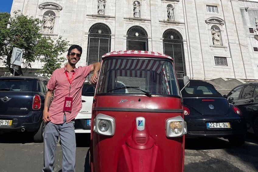 A man standing next to a red vehicle with a license plate, parked in front of a building with arched windows and statues. 2
