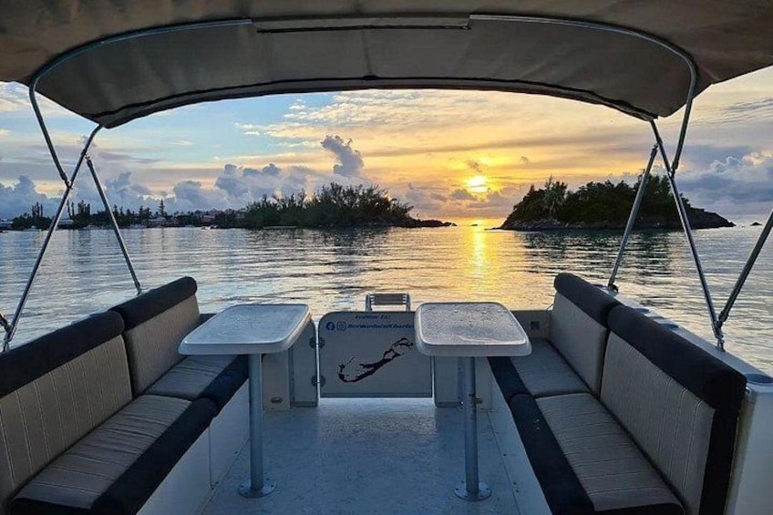 Een boot met twee tafels en een zonnescherm, met uitzicht op een zonsondergang boven het water en land in de verte. 5