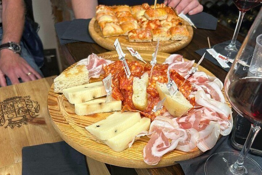 En skæringtallerken med forskellige slags kød og oste, en pizza i baggrunden og et glas rødvin på bordet. 3