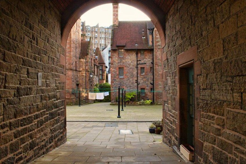 A stone archway leading to a courtyard with a paved path, brick buildings, and a potted plant. 3