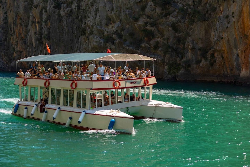 A boat with passengers, a canopy, and life rings, moving through turquoise water with a rocky cliff in the background. 3