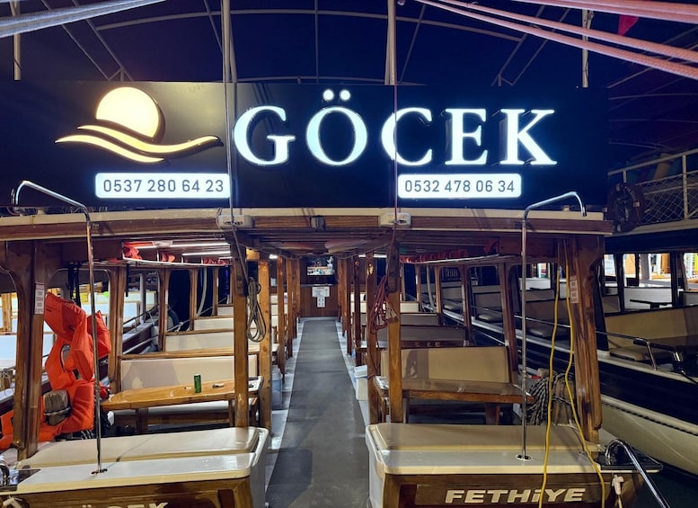 A ferry interior with seating and a sign reading 'GÖCEK' with contact numbers. 1