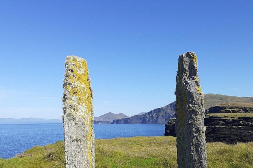 Two standing stones with lichen growth, a clear blue sky, and a distant view of a coastline with hills. 1