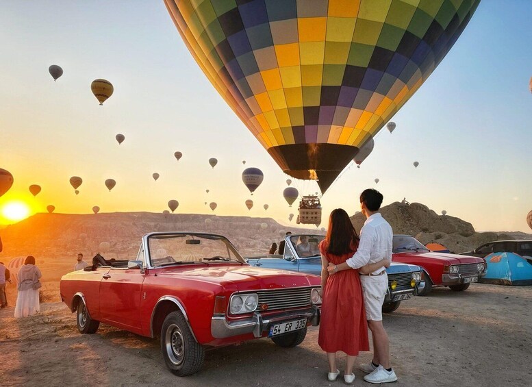 Una pareja de pie junto a un coche rojo antiguo, con globos aerostáticos al fondo. 5
