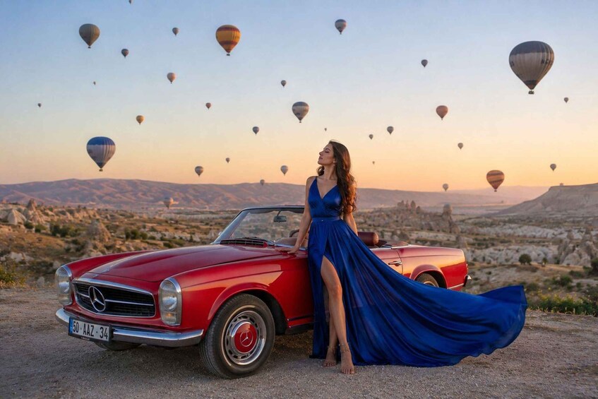 Una mujer con vestido azul posa junto a un coche descapotable rojo con placa de matrícula, rodeada de globos aerostáticos en un paisaje desértico. 2