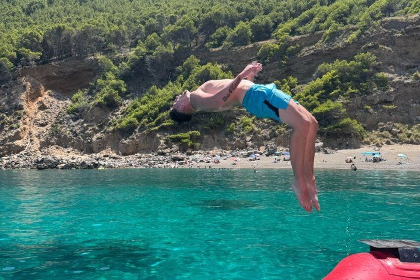 En person dykker ned i krystalklart turkis vand fra en klippe med en strand og parasoller i baggrunden. 2