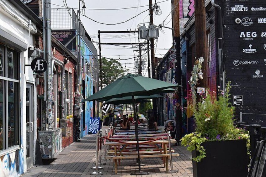 A narrow street with a green umbrella table, graffiti, and various storefronts. 3