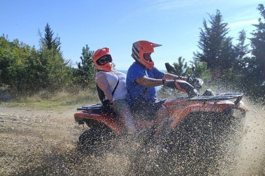 Two people riding an ATV on a dirt path surrounded by trees. 1