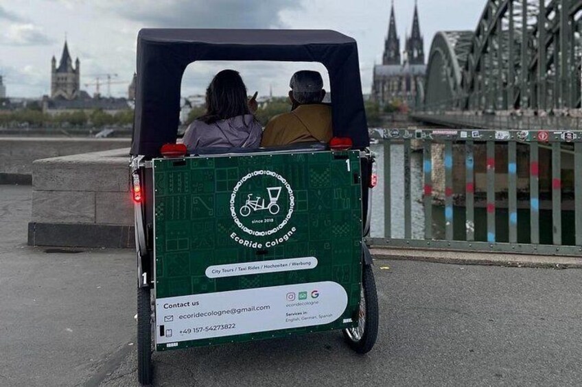 Een groene elektrische auto met een logo en contactgegevens op de achterkant, geparkeerd in de buurt van een brug, met een stadsgezicht op de achtergrond. 2