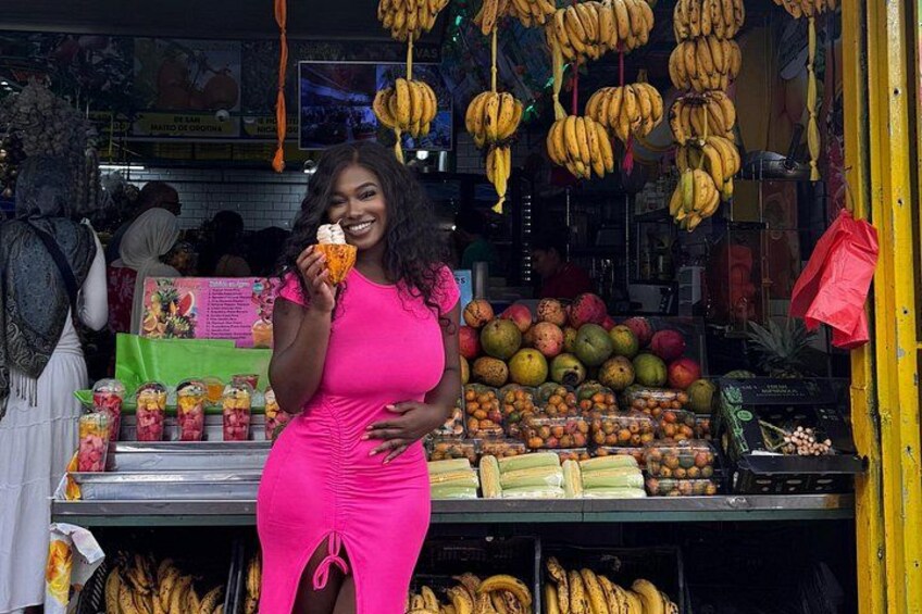 Eine Frau in einem leuchtend pinken Kleid steht vor einem Obststand, über dem verschiedene Früchte und Bananen hängen. 1