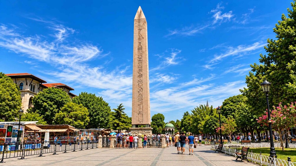 A tall obelisk with hieroglyphs, surrounded by a park with trees, benches, and a crowd of people. 4