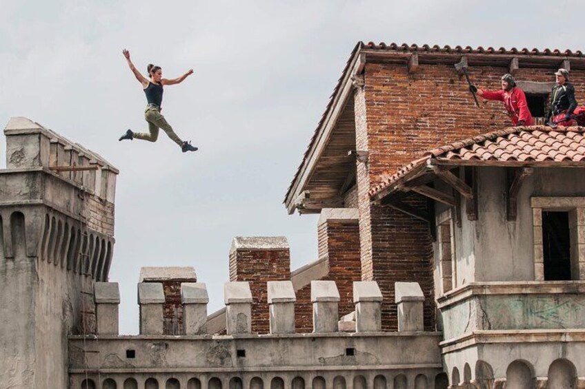 A person jumping off a building, another person on a roof, and a brick building with arches. 1