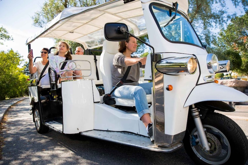 A white golf cart with passengers, driving on a paved road with trees in the background. 3