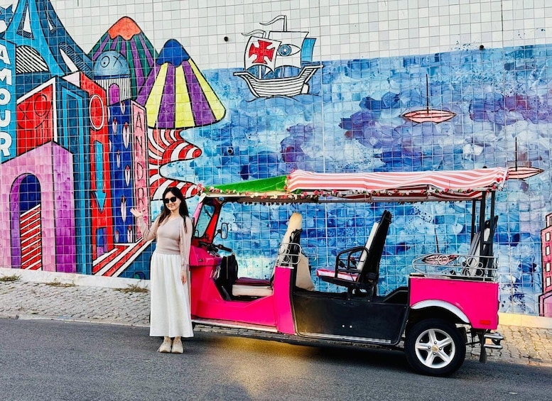 A pink tuk-tuk parked in front of a colorful mural with a ship and a striped umbrella. 2
