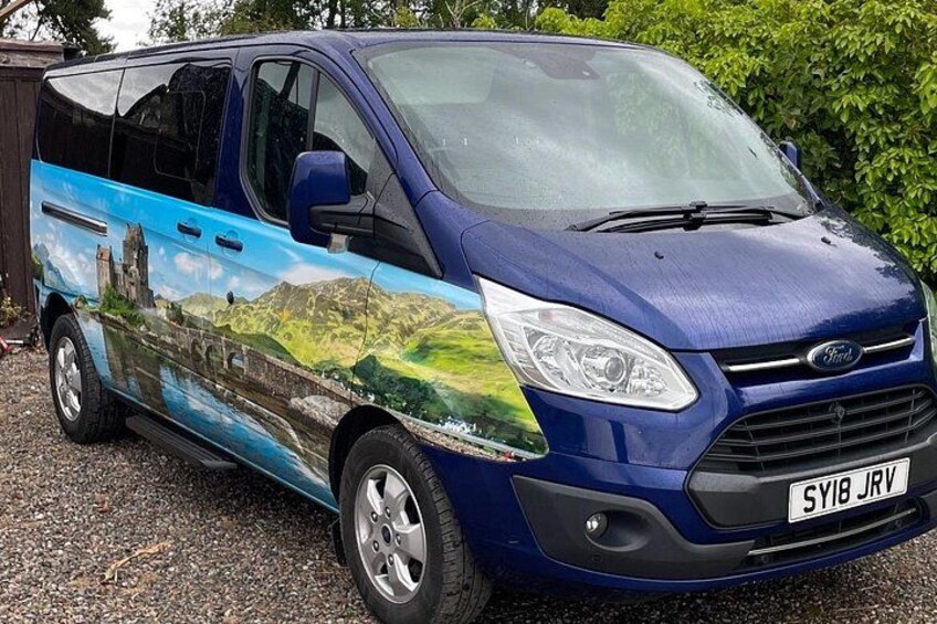 Een blauwe Ford bestelwagen met een decoratieve afbeelding van een landschap op de zijkant, geparkeerd op een grindpad. 2
