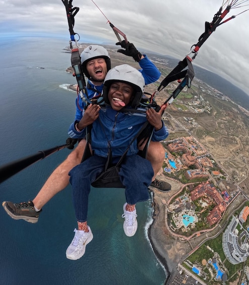 Two people paragliding over a coastal area with buildings and a beach visible below. 2