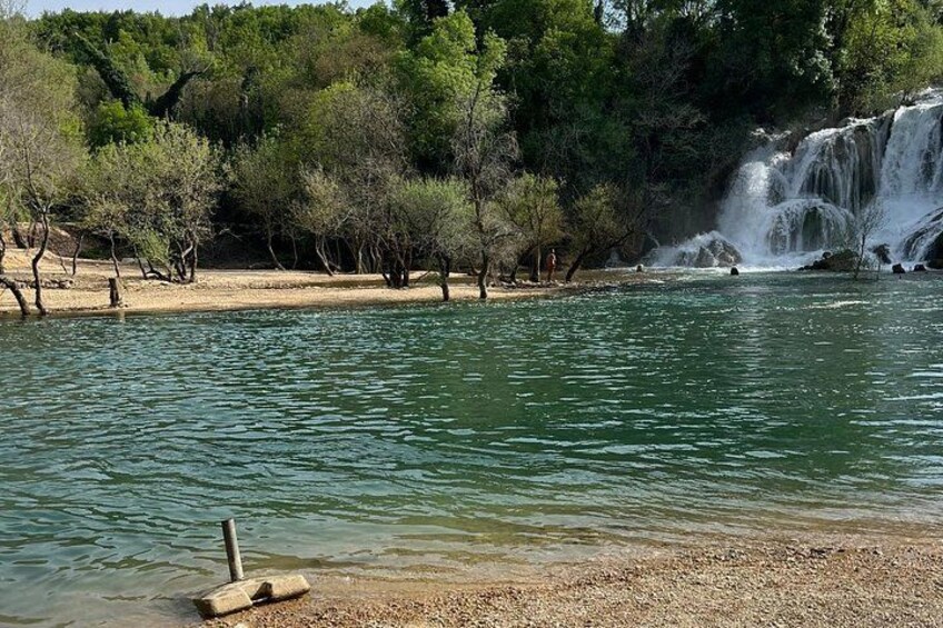 A calm lake with a waterfall, surrounded by trees and a pebbled shore. 3
