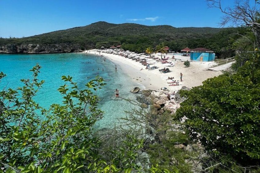 Ein Strand mit klarem türkisblauem Wasser, weißem Sand und einigen badenden Personen. Sonnenschirme und Liegestühle sind vorhanden, im Hintergrund sieht man ein kleines Gebäude. 4