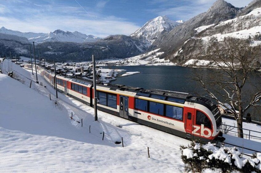A snow-covered train station with a train, mountains, and a lake in the background. 1