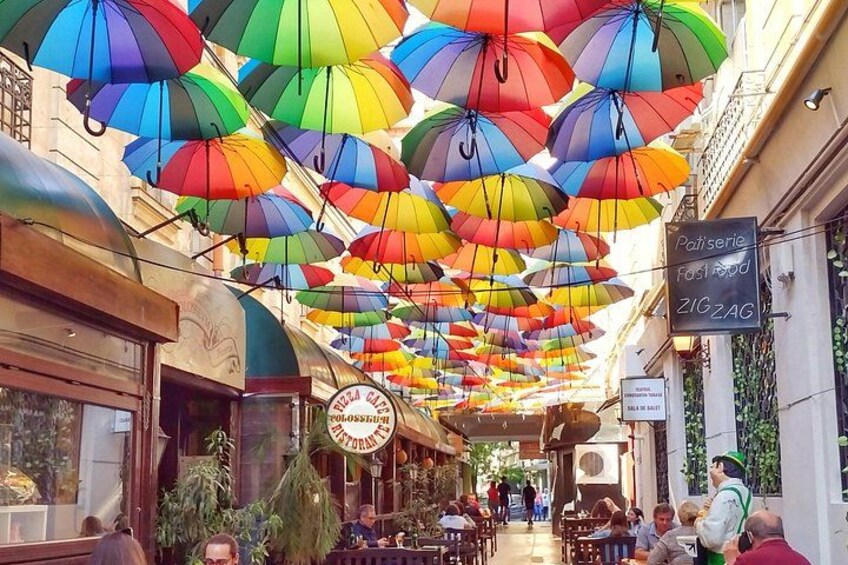 A street with colorful umbrellas hanging overhead, a café with outdoor seating, and a sign for a pastry shop. 4