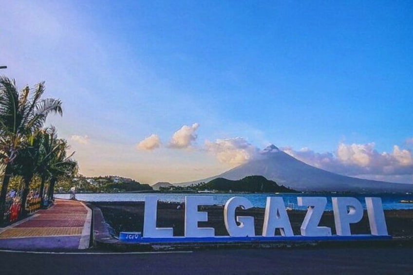 A sign with 'LEGAZPI' in large letters, palm trees, a body of water, and a mountain in the background. 1