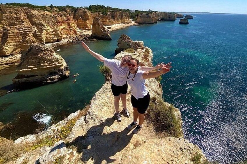 Dos personas de pie sobre un acantilado rocoso con vista a un paisaje costero con riscos y el mar. 4