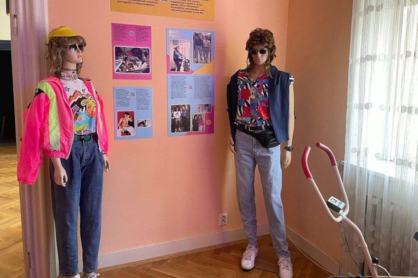 Zwei Schaufensterpuppen, eine in einer rosafarbenen Jacke und Jeans, die andere in einer blauen Jacke und Jeans, stehen in einem Raum mit einer rosafarbenen Wand und einem Fenster mit Vorhängen. 4