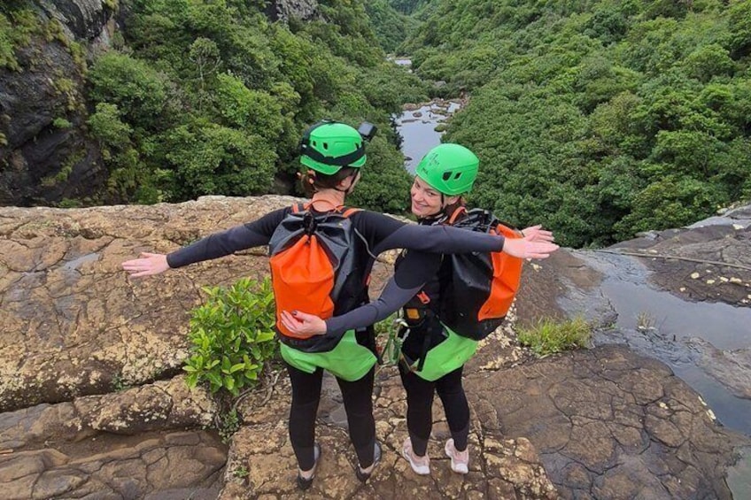 Deux personnes, portant des casques et des sacs à dos, se tiennent debout sur un promontoire rocheux, les bras tendus, et dominent une rivière ainsi qu’une végétation luxuriante. 3