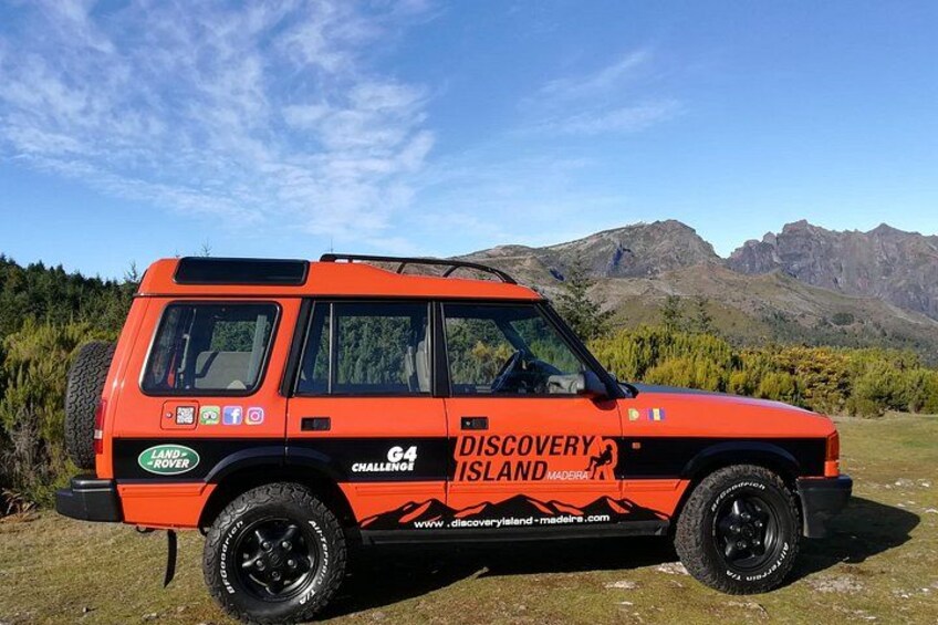 A red Land Rover Discovery with 'G4 Discovery Island' branding parked on grass with mountains in the background. 2