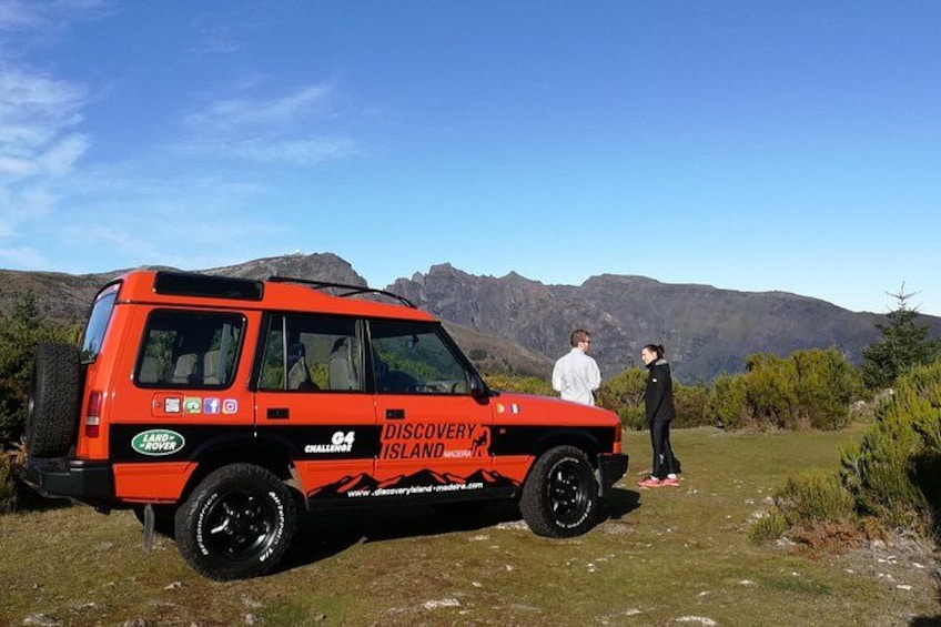 En röd Land Rover Discovery Islander med texten ”DISCOVERY ISLAND” och ”CHALLENGES” på sidan, parkerad på en gräsklädd åker med berg i bakgrunden. 5
