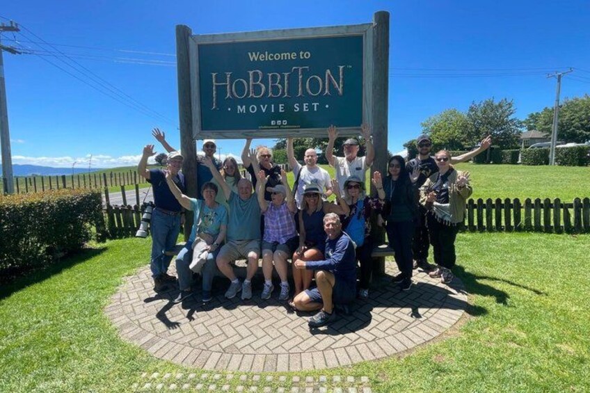 A group of people posing in front of a sign that reads 'Welcome to Hobbiton Movie Set.' 4
