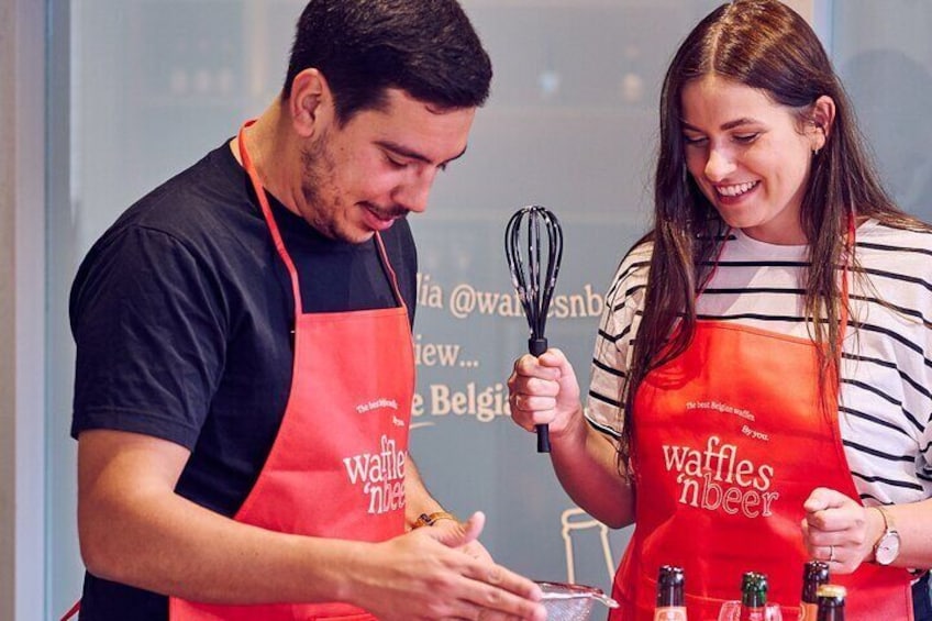 Two people wearing red aprons with the text "Waffles in Belgium" preparing food. 1
