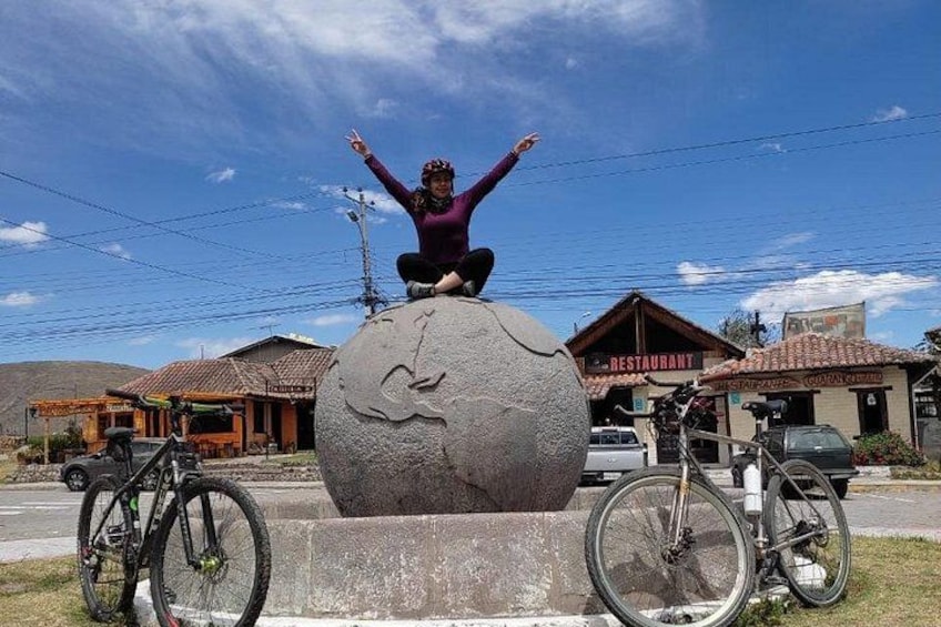 A person sitting on a stone sculpture with arms raised, a bicycle to the left, and buildings in the background. 1