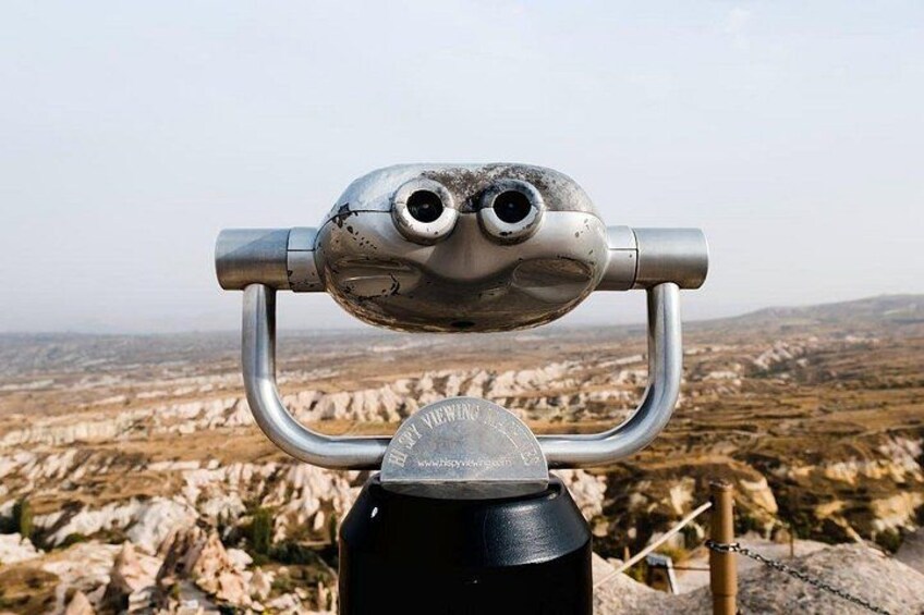 Unos binoculares de metal con estructura metálica, un objeto negro en primer plano y un paisaje rocoso al fondo. 5